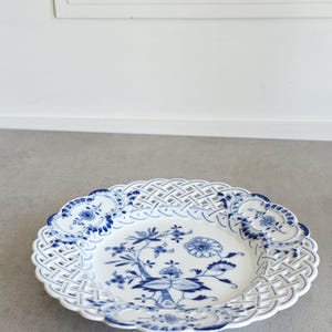 May include: A white porcelain plate featuring a blue floral pattern in the centre and a lattice-style openwork border. The rim is decorated with four blue floral medallions spaced evenly around the intricate woven-look edge of the dish.