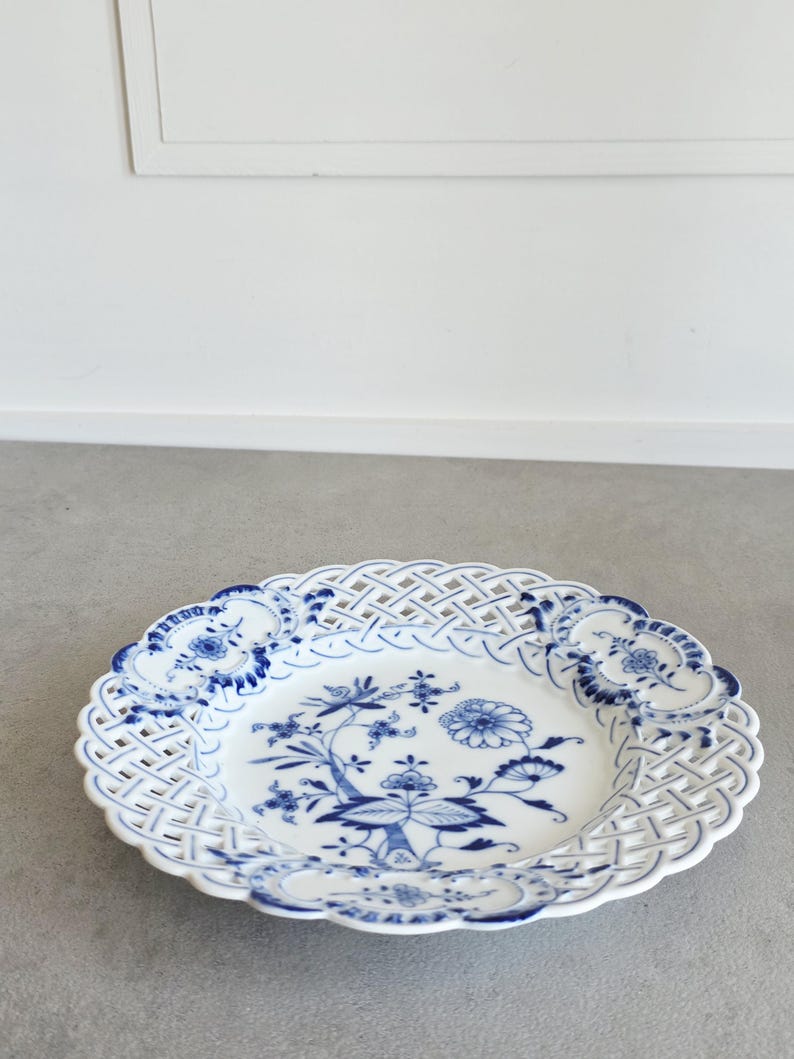 May include: A white porcelain plate featuring a blue floral pattern in the centre and a lattice-style openwork border. The rim is decorated with four blue floral medallions spaced evenly around the intricate woven-look edge of the dish.