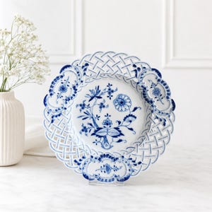 May include: A white porcelain plate featuring a blue floral pattern in the centre and a decorative lattice-style openwork border. The plate has scalloped edges with blue floral accents and is displayed on a clear stand.