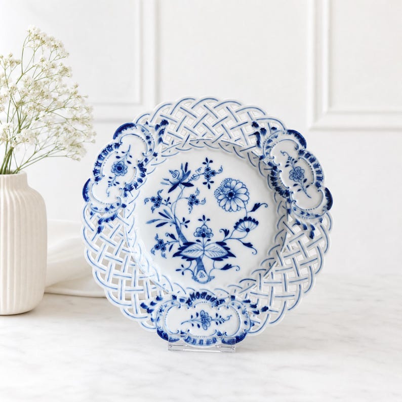 May include: A white porcelain plate featuring a blue floral pattern in the centre and a decorative lattice-style openwork border. The plate has scalloped edges with blue floral accents and is displayed on a clear stand.
