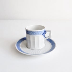 Op de afbeelding: Een wit porseleinen kop en schotel set met een blauwe patroonband rond de rand van de kop en de rand van de schotel. De kop heeft een verticaal geribbelde textuur en een vierkant handvat met een blauw bloemaccent aan de bovenzijde.