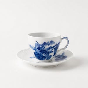 Pode incluir: Uma chávena de chá em porcelana branca com pires a condizer, apresentando um delicado padrão floral azul pintado no exterior da chávena e na superfície do pires. O conjunto tem uma subtil textura de cestaria em relevo no rebordo.