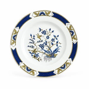 May include: A white ceramic dinner plate featuring a central illustration of a bird perched on a flowering branch with blue blossoms and green leaves. The rim is decorated with a repeating pattern of blue textured panels and floral motifs.