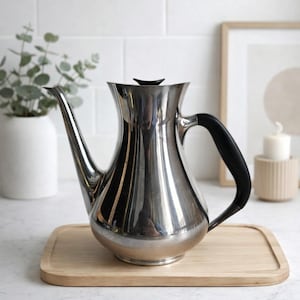 Puede incluir: Una cafetera plateada brillante con un asa y tapa negras. La cafetera tiene un pico largo y curvado y está sobre una bandeja de madera clara. El fondo incluye una pared de azulejos blancos, un cuadro enmarcado y un jarrón blanco con vegetación.