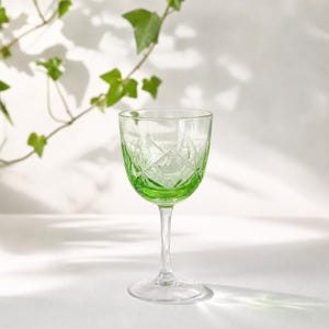 Può includere: Un bicchiere da vino tinto di verde con stelo e base trasparenti. Il bicchiere presenta un motivo a taglio diamante sulla coppa. Lo sfondo è bianco con ombre e un vitigno.