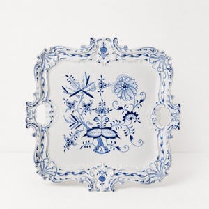 Może przedstawiać: Kwadratowa taca do serwowania z białej porcelany z misternym niebieskim wzorem kwiatowym pośrodku oraz ozdobnymi, falistymi krawędziami z niebieskimi detalami. Taca posiada dwa zintegrowane uchwyty boczne i symetryczny wzór obrzeża.