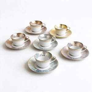 Puede incluir: Un juego de seis tazas de té y platillos de porcelana blanca, cada uno con un borde texturizado de metal dorado o plateado y un platillo a juego. Las tazas tienen un interior blanco liso y un asa lateral, dispuestas en círculo.