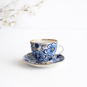 May include: A white teacup and saucer set with a blue floral pattern and gold trim. The teacup has a handle and sits on a matching saucer. The floral design features blue flowers and gold accents.