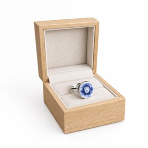 Puede incluir: Un anillo de plata con una incrustación circular de patrón floral azul y blanco, presentado dentro de una caja de madera clara abierta con un forro interior de terciopelo beige. El anillo tiene una banda de metal pulido y una cara redonda.