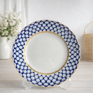 Peut inclure: Assiette en porcelaine blanche avec un motif en treillis bleu et or sur le bord. L'assiette a un bord festonné et une bordure dorée. L'assiette est présentée sur un support transparent.