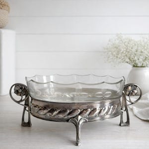 Peut inclure: Un saladier en verre transparent avec un bord festonné est posé sur un support en métal argenté. Le support présente un motif décoratif en forme de corde autour de la base et des motifs floraux sur les poignées. Le bol repose sur trois pieds.