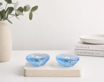 May include: Two small, circular, light blue glass candle holders with a textured, rippled surface. Each piece features a central indentation designed to hold a candle, showcasing a translucent finish that catches the light.