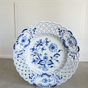 May include: A white porcelain plate featuring a blue floral pattern in the centre and a decorative lattice-style openwork border. The rim is accented with three distinct blue floral cartouches, creating a detailed and intricate circular design.
