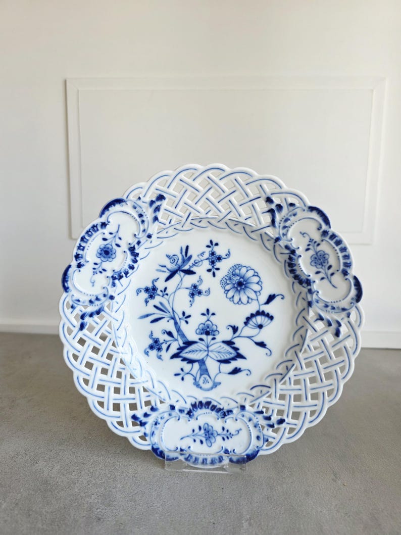 May include: A white porcelain plate featuring a blue floral pattern in the centre and a decorative lattice-style openwork border. The rim is accented with three distinct blue floral cartouches, creating a detailed and intricate circular design.