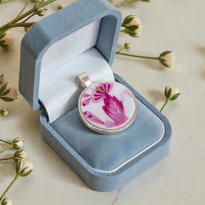 May include: A round pendant with a floral design in shades of pink and white, set in a silver-colored metal. The pendant rests in a light blue velvet jewelry box. Delicate white flowers and greenery are arranged around the box.