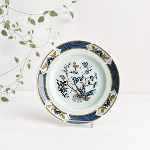 May include: A white ceramic plate with a blue and gold border. The plate features a central design of a bird and floral branches in shades of blue and brown. The plate is displayed on a clear stand.