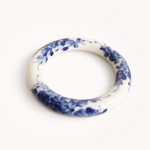 May include: A circular bangle bracelet made of white ceramic material. The surface of the bracelet is decorated with a continuous hand-painted pattern of blue floral motifs and delicate leafy vines that wrap around the entire circumference.