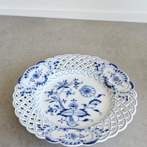 May include: A white porcelain plate featuring a blue floral pattern in the centre and a decorative lattice-style openwork border. The rim is accented with four distinct blue floral motifs, creating a detailed and intricate design on the dish.