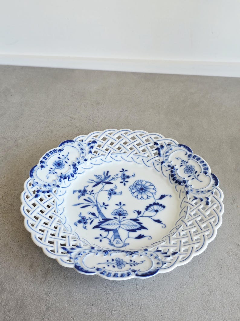 May include: A white porcelain plate featuring a blue floral pattern in the centre and a decorative lattice-style openwork border. The rim is accented with four distinct blue floral motifs, creating a detailed and intricate design on the dish.