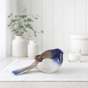 May include: A ceramic bird ornament with a blue head and wings, brown back, and white chest. The bird is positioned on a white surface. The figurine has a long tail and is a decorative item.