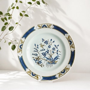 Puede incluir: Un plato de cerámica blanca con un borde geométrico azul y dorado. El centro presenta una ilustración detallada de un pájaro y ramas florales en tonos azules, marrones y verdes. El plato se exhibe sobre un soporte transparente.