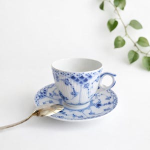 Può includere: Un set di tazza e piattino bianchi con un motivo floreale blu e un cucchiaino d'argento. La tazza ha un manico ed è posizionata su un piattino abbinato. Il design presenta intricati dettagli blu, creando un'estetica elegante.
