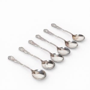 Könnte beinhalten: Ein Set aus sechs silberfarbenen Metalllöffeln, die in einer diagonalen Linie angeordnet sind. Jeder Löffel hat eine polierte ovale Laffe und einen schlanken Stiel, der mit einem komplexen, erhabenen Blumenmuster an der Spitze verziert ist.