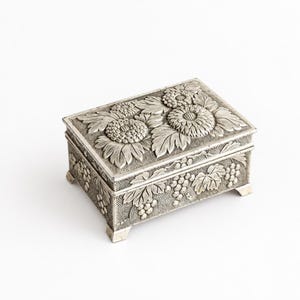 Pode incluir: Uma caixa retangular de metal prateado com um design floral em relevo na tampa, apresentando grandes flores semelhantes a crisântemos e folhas. As laterais da caixa são decoradas com um padrão em relevo de cachos de uvas e folhas.