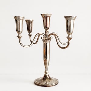 May include: A silver-toned metal candelabra featuring four candle holders arranged on curved arms extending from a central pillar. The structure stands on a circular base, with a polished, reflective surface and a symmetrical design throughout.