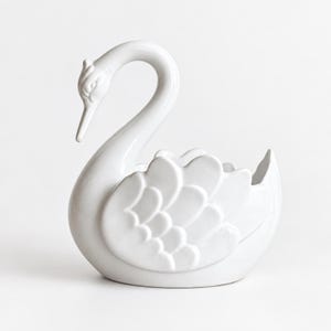 Może przedstawiać: Biała ceramiczna miska w kształcie łabędzia z błyszczącym wykończeniem. Łabędź ma długą, wygiętą szyję i głowę lekko zwróconą w bok. Korpus został zaprojektowany z rzeźbionymi, nakładającymi się na siebie piórami skrzydeł, które tworzą misę.