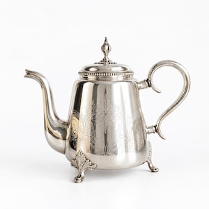 Op de afbeelding: Een gepolijste zilveren theepot met een sierlijke bloemengravure op de behuizing, een gebogen handvat, een lange tuit en een deksel met een decoratieve knop. De theepot staat op drie gedetailleerde pootjes en heeft een parelrand rond de deksel.