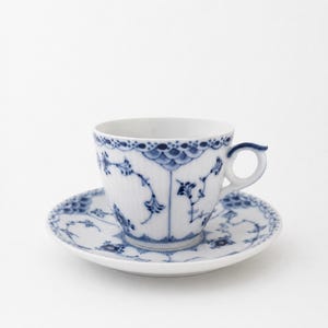 May include: A white porcelain teacup and saucer set featuring a delicate blue floral pattern. The cup has a ribbed texture, a curved handle, and a scalloped rim design. The saucer matches the cup with intricate blue botanical motifs.