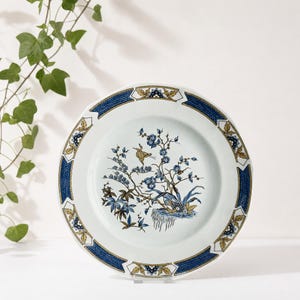 May include: White ceramic plate with a blue and gold border. The center features a detailed illustration of a bird and floral branches in shades of blue, brown, and gold. The plate is displayed on a clear stand.