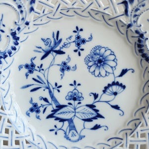 May include: A white porcelain plate featuring a blue floral pattern in the centre and a decorative lattice-style openwork border. The intricate design includes various stylised flowers, leaves, and stems painted in shades of cobalt blue.