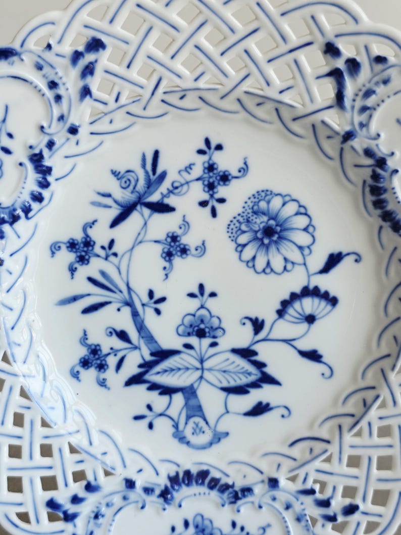 May include: A white porcelain plate featuring a blue floral pattern in the centre and a decorative lattice-style openwork border. The intricate design includes various stylised flowers, leaves, and stems painted in shades of cobalt blue.