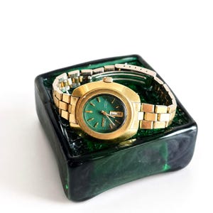 May include: A gold-tone Seiko wristwatch with a round green dial, gold-tone hour markers, and a day-date window at the three o'clock position. The watch features a metallic link bracelet and is resting inside a square, dark green glass dish.