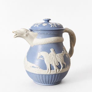 May include: A light blue ceramic jug with white relief detailing. The spout is shaped like a dolphin head, and the handle resembles a coiled tail. The body features a white embossed scene of a person standing next to a horse.