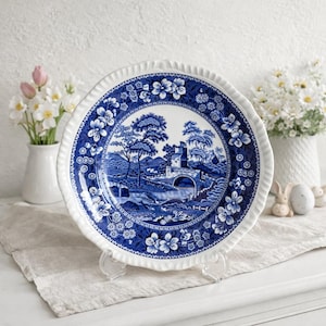 Op de afbeelding: Een blauw en wit decoratief bord met een landschapsscène. Het bord heeft een geschulpte rand met bloemendetails. De centrale afbeelding toont een brug, bomen en een gebouw. Het bord wordt op een transparante standaard gepresenteerd.