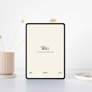 May include: A digital tablet with a black border displays the text "weekly RESET" and "A Gentle Mental Health Journal" on a cream-coloured background. The words "calm", "reflect", and "reset" are at the bottom. A beige vase with dried flowers and a white plate are in the background.