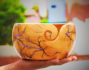 May include: A wooden yarn bowl with a purple and black swirl design. The bowl has a unique shape with a hole in the side for yarn to pass through.