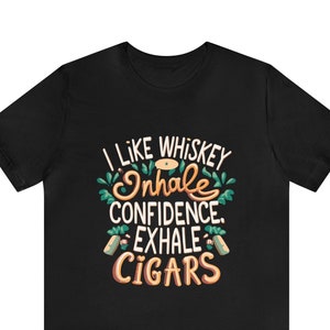 May include: Black t-shirt with the text "I LIKE WHISKEY INHALE CONFIDENCE EXHALE CIGARS" in a decorative font. The text is in white, green, and orange. The shirt is a short-sleeved crew neck.