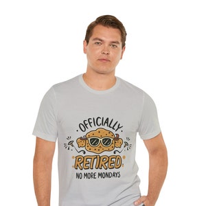 May include: Light gray t-shirt with the text "OFFICIALLY RETIRED. NO MORE MONDAYS" and a cartoon image of a cookie wearing sunglasses. The shirt is made of a soft material and has short sleeves.