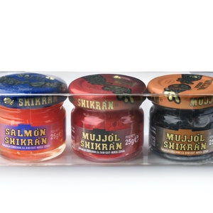 Könnte beinhalten: Eine transparente Kunststoffverpackung mit drei kleinen Gläsern mit Fischrogen. Die Gläser haben farbige Deckel und Etiketten mit der Aufschrift "Salmon Shikran", "Mujjol Shikran" und "25g". Der Rogen in den Gläsern ist orange, rot und schwarz.