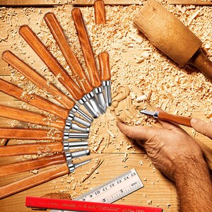 May include: A set of 15 wood carving tools with wooden handles and metal blades. The tools are arranged in a fan shape on a wooden surface. A hand is holding a small carving tool. The tools are used for woodworking and crafts.