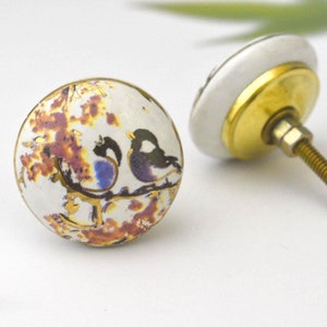 Peut inclure: Deux boutons de porte en céramique avec une bordure dorée. Un bouton présente un fond blanc avec un motif floral brun et or et deux oiseaux. L'autre bouton est blanc uni.