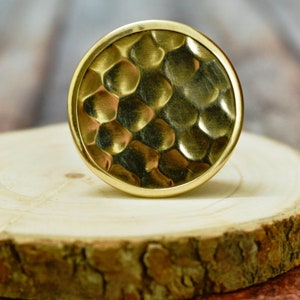 May include: A round, gold-colored metal knob with a hammered texture. The knob is sitting on a wooden surface.