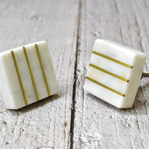 Puede incluir: Dos pomos cuadrados blancos para armarios con rayas doradas. Los pomos tienen un tornillo en la parte posterior para sujetarlos a una puerta de armario.