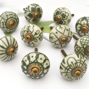 Op de afbeelding: Een verzameling van tien decoratieve keramische kastknoppen. Elke knop heeft een crèmekleurige basis met donkergroene bloempatronen en een messingkleurige metalen basis. De knoppen zijn gerangschikt op een witte ondergrond.