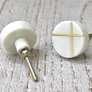 Op de afbeelding: Twee witte marmeren kastknoppen met gouden kruisontwerpen. De knoppen zijn rond en hebben een schroef aan de achterkant.