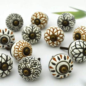 Op de afbeelding: Een verzameling decoratieve keramische kastknoppen. De knoppen hebben bloempatronen in wit, zwart en bruin, met messingkleurige hardware. De ronde knoppen hebben een klassiek, elegant ontwerp.